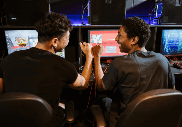 Dua gamer sedang fokus bermain di depan banyak monitor, menunjukkan intensitas dalam tren industri game.